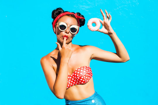 Beautiful, funny, cheerful,  tanned girl eats a sweet donut glaze, licks his fingers, dressed in a bright bikini, sunglasses, hairstyle with horns, makeup, lovely emotions, blue wall, perfect body