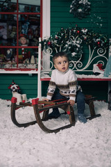 Obraz premium Little boy enjoying sleigh ride. Child sledding on sledge. Green wall with a window, bench is decorated with a Christmas wreath. Snow flakes. Studio shoot