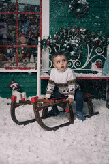 Little boy enjoying sleigh ride. Child sledding on sledge. Green wall with a window, bench is decorated with a Christmas wreath. Snow flakes. Studio shoot