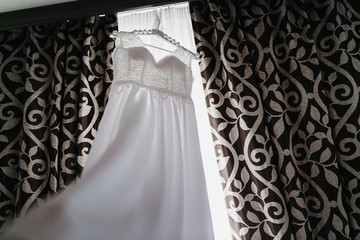 White Wedding dress hang on a shoulders near window, before ceremony