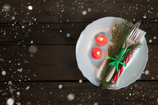 Country style christmas decoration. White empty plate and two red lighted candles with fir tree, candy, fork and knife tied with a green ribbon on old wooden brown background.