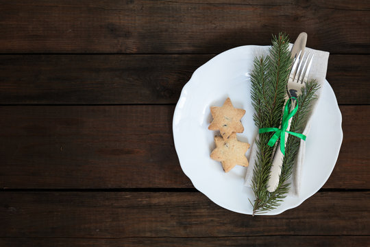 Country style christmas decoration. White empty plate and Christmas cookies with fir tree, candy, fork and knife tied with a green ribbon on old wooden brown background.