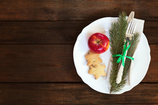 Country style christmas decoration. White empty plate, red apple and Christmas cookies with fir tree, candy, fork and knife tied with a green ribbon on old wooden brown background.