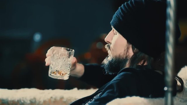 Young bearded man drink alcohol at bar HD slow motion video. Guy in hat with whiskey glass relaxing in club, sitting in armchair alone