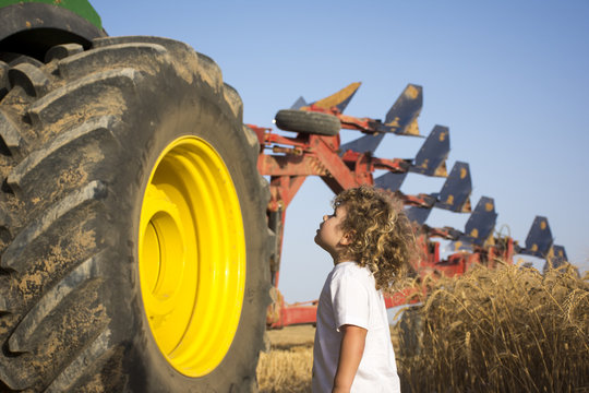 A Toddler looks up at the wheel of a big tractor with plough on the back in a wheat field