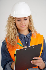 Young female architect with blonde, curly hair and wearing safety vest and white hardhat taking notes
