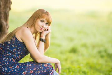 Beautiful young girl sitting against a tree on the background of blurred green grass and looks into the distance