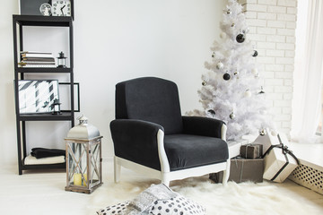 Modern stylish Christmas decor of white living room with brick wall and wood floor. Black armchair, bookshelf, lantern, white plastic tree decorated with dark toys and presents in wrapping paper.