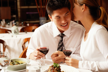 Couple in restaurant, sitting side by side, woman whispering into man's ear