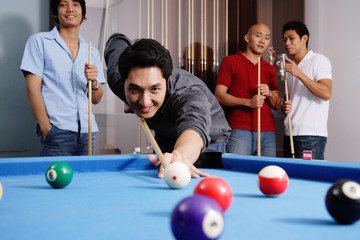 Man aiming pool cue at ball, other men watching in the background