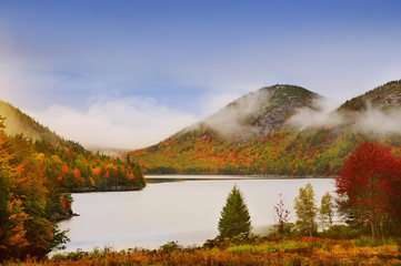 Lake in the mountains, colors around autumn trees.   Acadia National Park. Maine. USA. Cloudy gloomy weather, fog and clouds over the mountains in the distance. 
