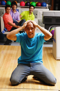 Man kneeling in bowling alley, hands on head, frowning, people in the background watching