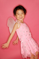 Young girl in pink dress with artificial wings, smiling at camera, hands stretched behind her