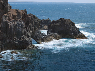 Ocean waves breaking on the rocky coast of hardened lava with caverns and cavities. Mountains and volcanoes on the horizon. Lanzarote, Canary Islands, Spain