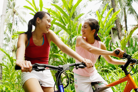 Two girls riding bikes through a park, looking at each other