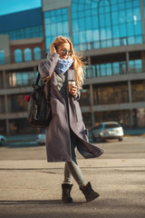 Fototapeta premium Great weather today for walk. Beautiful young blonde woman holding coffee cup and walking outdoors with smile on her face. Sunny day, happiness, coffee break, enjoying.