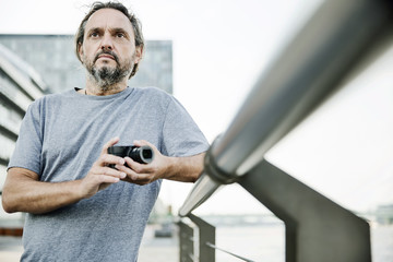 Mann steht an einem Geländer mit einer digitalen Kompaktkamera in der Hand, Portrait, 