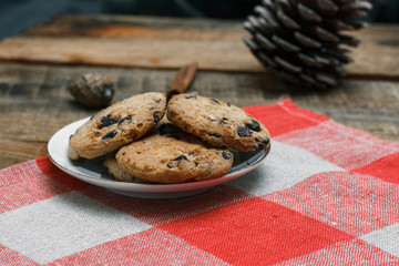 Chocolate cookies in a white plate on a checkered tablecloth on wooden background. Near cones and cinnamon stick, roughly. The theme of the new year holiday