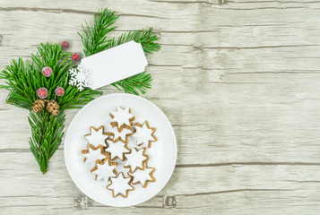 Obraz na płótnie Canvas Kleiner runder weißer Teller mit Weihnachtsplätzchen, Zimtsternen, einem Tannenzweig und gefrorenen roten Beeren und einem weißen Schild, das beschriftet werden kann, Untergrund Holz, Textfreiraum