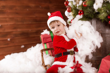 Small child (boy) in the costume of Santa Claus throws white snow up near the Christmas tree with gifts
