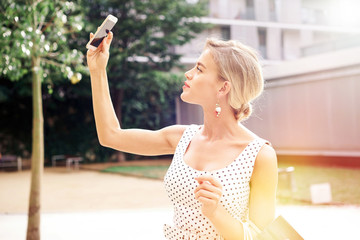 A waist up photo with flare light of a beautiful caucasian female wearing white summer dress and taking photo by a camera of a smartphone while standing with the bags on a sunny street.