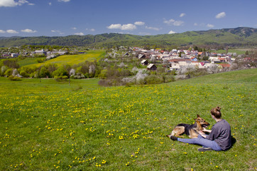 Girl and dog are sitting on spring meadow and they are looking to wonderfull village which is full of colors and pleasure afternoon light. They love each other and they are best friends