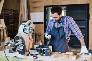 carpenter texting someone on his smart phone near circular saw in a dusty workshop