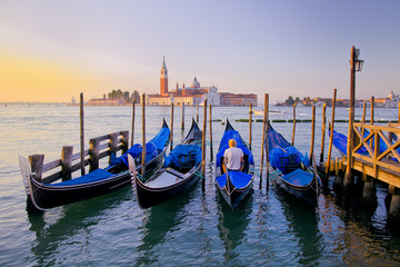 Young man in white clothes sitting in front of Grand Canal in Venice with gondolas against San Giorgio Maggiore church in Italy. Scenery from summer vacation with beautiful colorful morning light.
