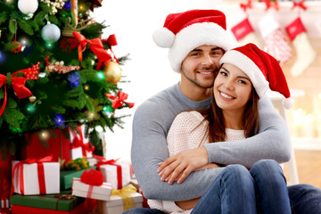 Young couple in Santa Claus hats sitting near Christmas tree indoors