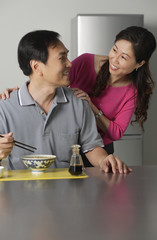 Mature man in kitchen, eating a bowl of noodles, turning to look at woman standing behind him