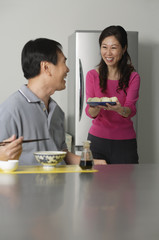 Mature couple in kitchen, man sitting in front of a bowl of noodles, woman behind him holding a plate of food