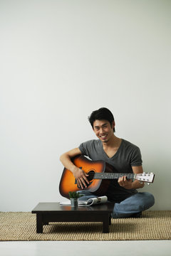 Young man sitting on floor playing guitar smiling at camera