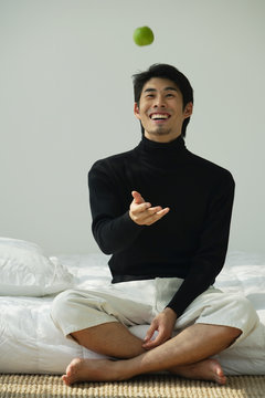 Young man sitting on bed, throwing apple in the air