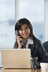 A woman looks at the camera and talks on the phone as she sits at her desk