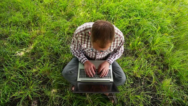 Young man in glasses sitting on the grass and typing on the lapt