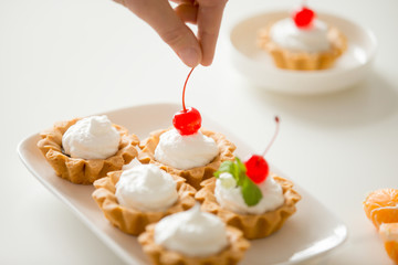 Close up of hand decorating colorful mini fruit and cream tarts with red cherry, set in a row on a white table