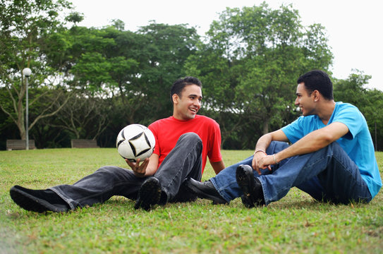 Two men sitting on grass, talking, one holding soccer ball