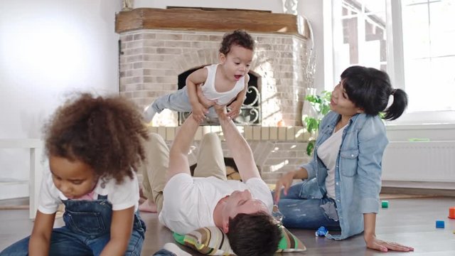 Parents spending time with kids in living room, happy dad lying on the floor, lifting his baby son, mom holding his hand carefully as cute curly daughter sitting and playing next to them 