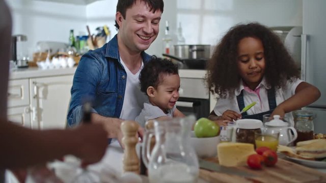 Rack focus of moms hand whisking while father sitting at table with his little son on lap and cute curly daughter sitting next to them, kids playing with kitchen stuff happily