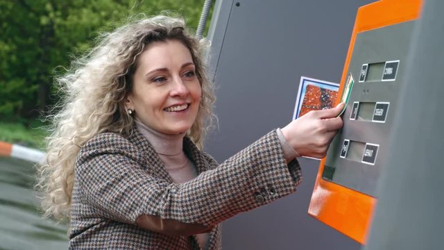 Slow motion of attractive middle-aged curly blonde woman in casual wear jumping joyfully on corner of kiosk in order to pay for petrol in filling station