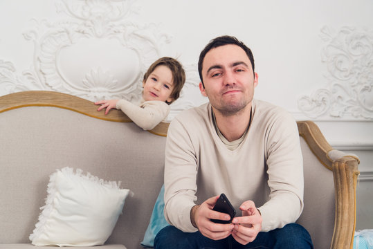 Tired Frustrated Father Sitting On A Sofa Using Phone And Son Who Wants To Play With Him