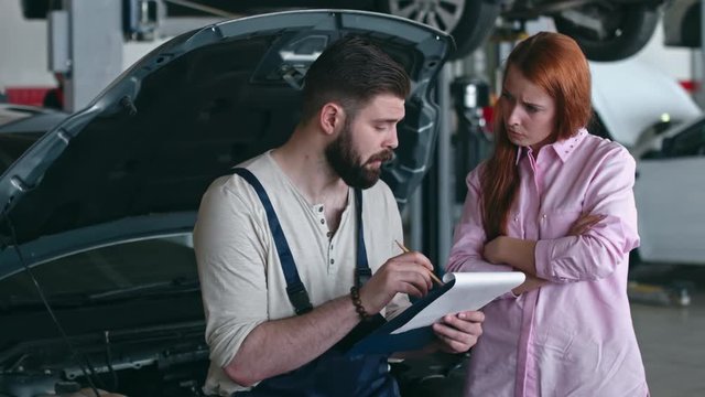Car technician reading document and explaining something to young woman in auto service