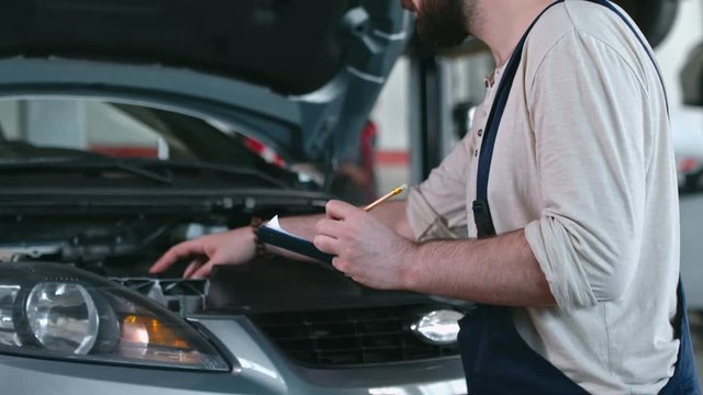 Auto mechanic in professional uniform checking car engine and making some notes