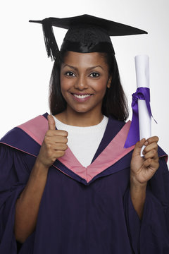 Young woman wearing cap and gown and holding diploma giving thumbs up sign