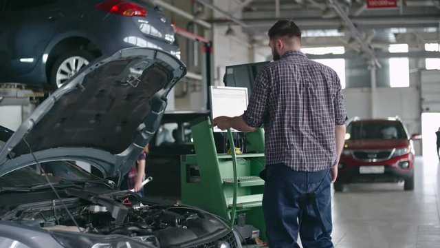 Auto repairman examining details under the hood of car and using computer to check breakdown