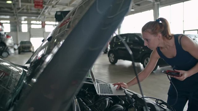 Female mechanic in professional uniform using laptop and special device for car diagnostics