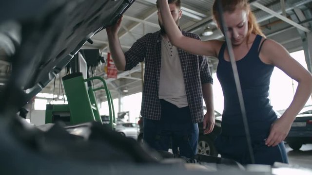 Man and woman in mechanic uniform checking details under car hood and fixing the breakdown