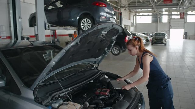 Woman walking to car in the repair garage and using laptop for auto diagnostics