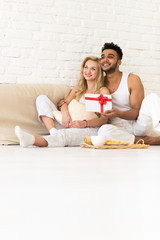 Obraz na płótnie Canvas Young Couple Sit On Pillows Floor, Happy Smile Hispanic Man Woman Hold Present Envelope With Ribbon Lovers In Bedroom Over White Brick Wall