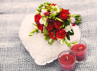 Beautiful romantic decor in red colors for wedding or dating of young couple. Small bouquet of flowers on white pillow in shape of heart and two red candles in glass holders on picnic blanket.